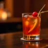 A classic Manhattan cocktail garnished with a maraschino cherry and citrus peel in an elegant, dimly lit bar.