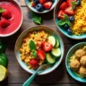 Colorful gluten-free vegan dishes including quinoa Buddha bowl, tofu scramble, smoothie bowl, zucchini noodles, and energy bites arranged on a rustic wooden table.